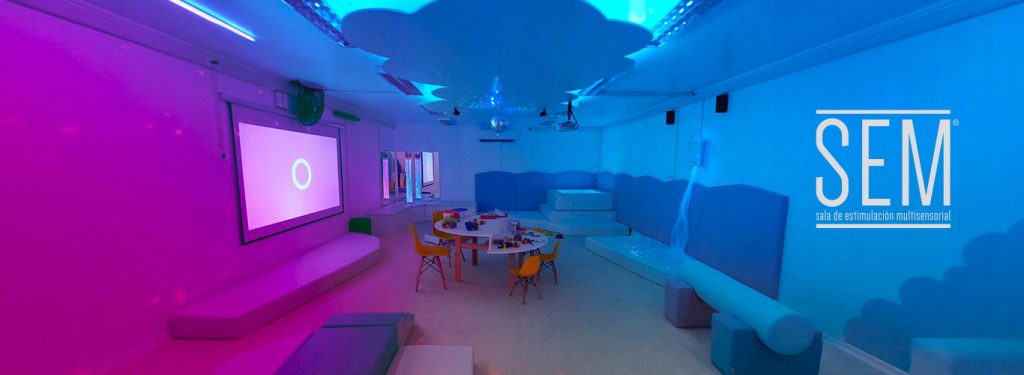 Vista panorámica de una sala multisensorial. En la imagen se aprecian los elementos: mesas de trabajo grupal, juegos de espuma, piscina de pelota retroiluminada, proyector con telón, tubos de burbujas. bola con espejos, nube en el techo con luces. el ambiente destaca los colores azul y lila. Se destaca el logo "SEM, SALA DE ESTIMULACIÓN MULTISENSORIAL"
