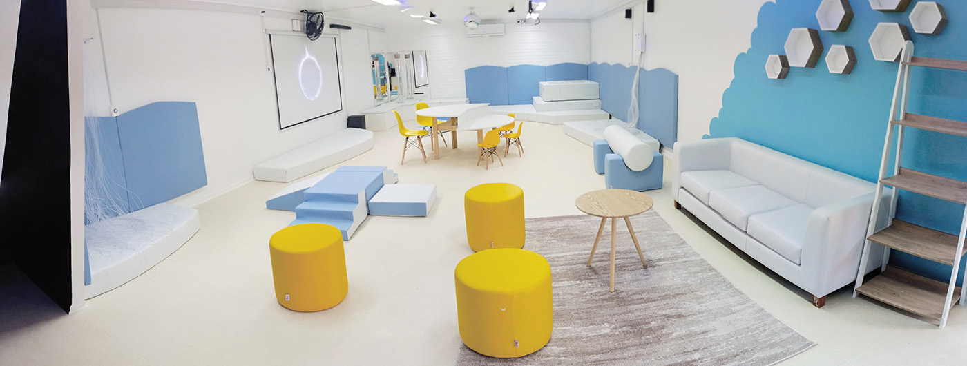 Imagen panorámica de una Sala de Estimulación Multisensorial SEM con todas las luces frías encendidas. Destaca la piscina con pelotas, tubo de burbujas, proyector en el muro, bola de espejos, puf amarillos, alfombra con mesa pequeña, juegos de espuma para el desarrollo de la motricidad, mesas de trabajo, sillones, ventilador, fibra óptica.