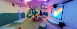 Vista panorámica de una sala multisensorial. En la imagen se aprecian los elementos: mesas de trabajo grupal, juegos de espuma, piscina de pelota retroiluminada, proyector, telón en el que se aprecia un arcoíris de colores, tubos de burbujas. bola con espejos, nube en el techo con luces.