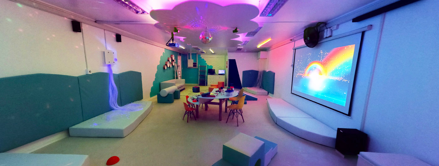 Vista panorámica de una sala multisensorial. En la imagen se aprecian los elementos: mesas de trabajo grupal, juegos de espuma, piscina de pelota retroiluminada, proyector, telón en el que se aprecia un arcoíris de colores, tubos de burbujas. bola con espejos, nube en el techo con luces.