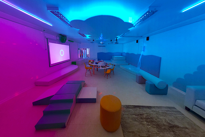 Vista panorámica de una sala multisensorial. En la imagen se aprecian los elementos: mesas de trabajo grupal, juegos de espuma, piscina de pelota retroiluminada, proyector con telón, tubos de burbujas. bola con espejos, nube en el techo con luces. el ambiente destaca los colores azul y lila. Se destaca el logo "SEM, SALA DE ESTIMULACIÓN MULTISENSORIAL"