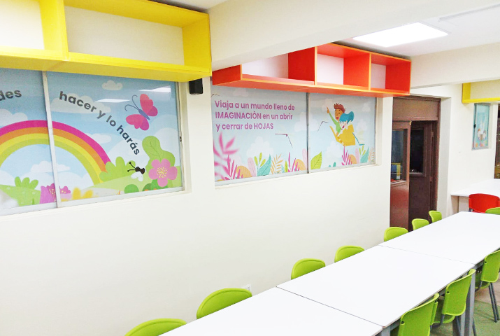 Vista de una parte de una biblioteca. En la imagen aparece coloridas repisas, mesas de estudio y gráficas en las ventanas con frases motivadoras que fomentan la lectura.